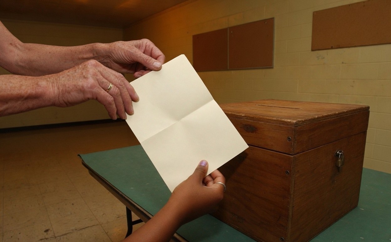 Illustrative scene of two people holding a folded blank ballot beside a wooden ballot box